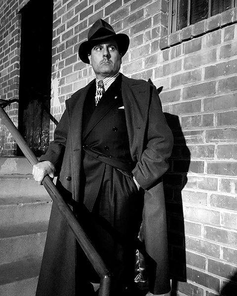 Jacob Caputo, owner and operator of Haunted Whittier Tours, stands on stairs looking into the night pensively. He is impeccably dressed in a 1940s suit and overcoat, wearing a fedora hat in this black and white photo.