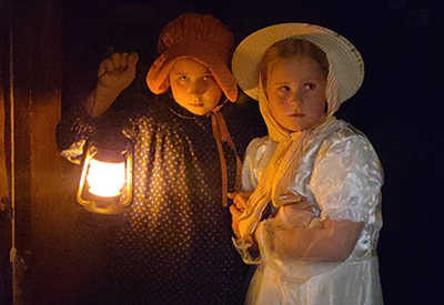 Young twin girls, dressed in frontier dresses and bonnets, hold a lantern in the night, looking apprehensive.